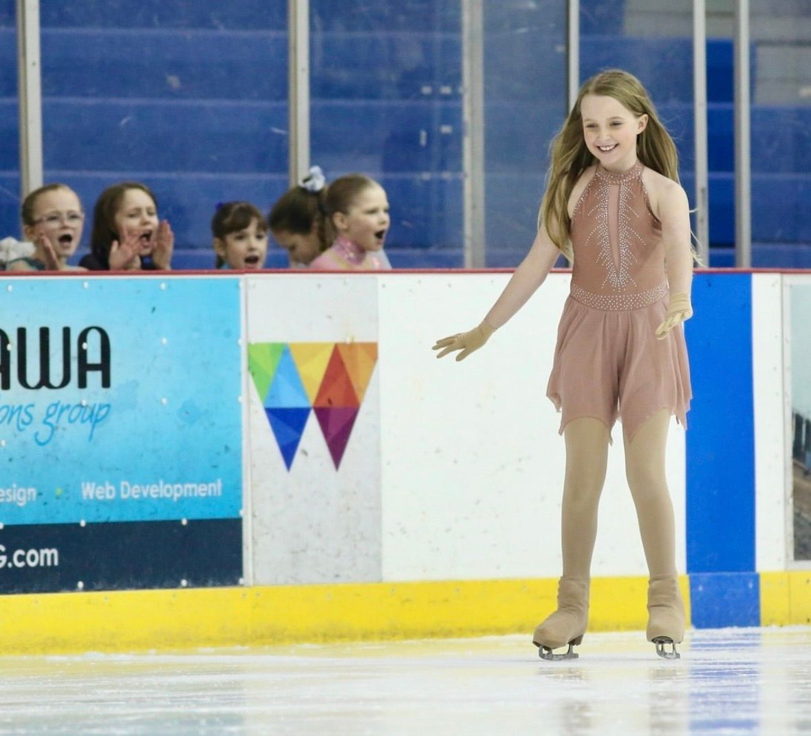 Young skater in pink dress, performing on ice. Audience watches in awe from the barrier.