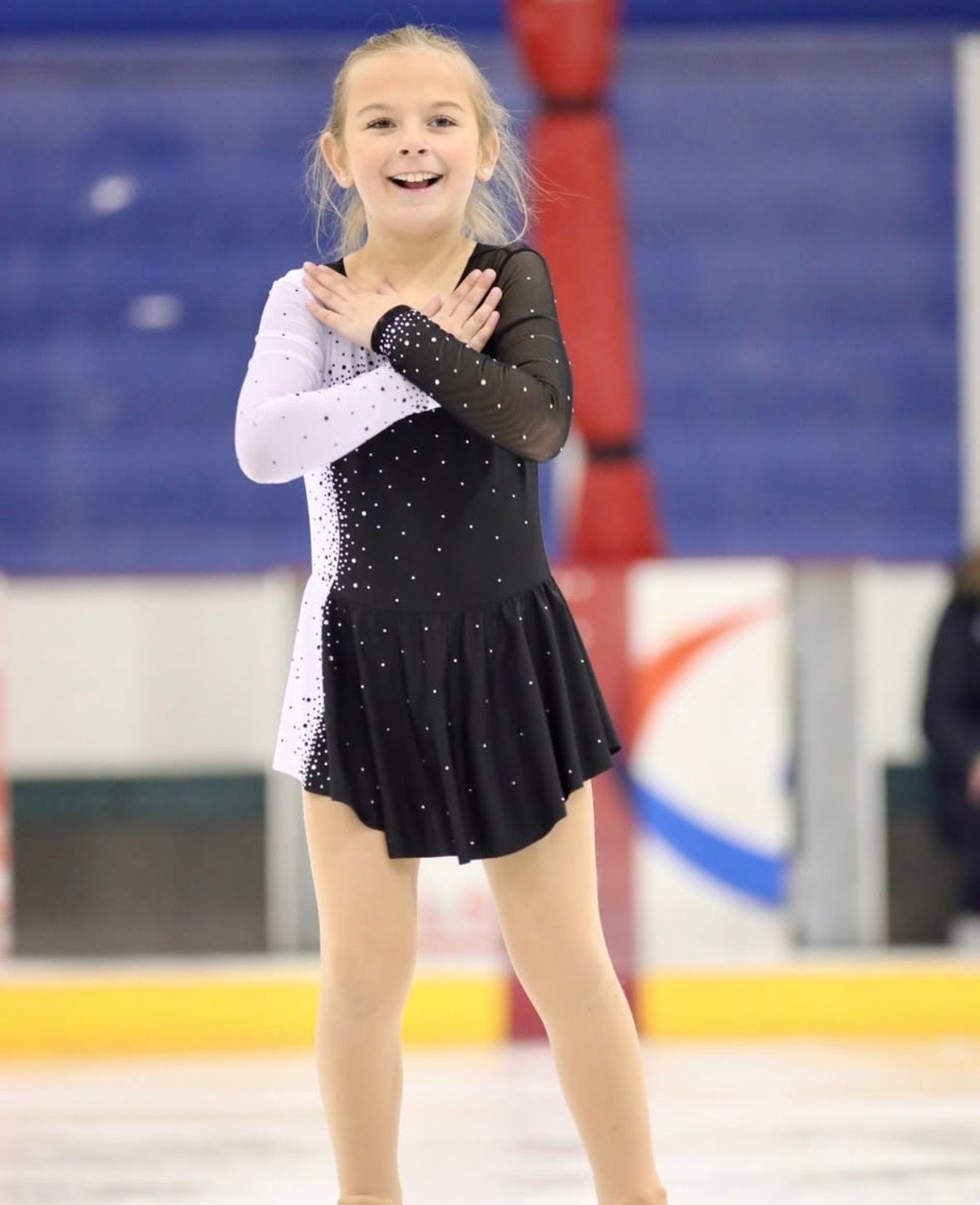 Young figure skater in a black and white dress, arms crossed, smiling on ice rink.