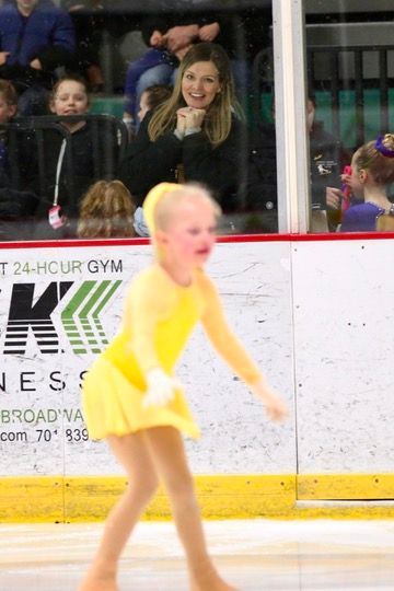 Young figure skater in yellow dress performs, crowd watches from rink side.