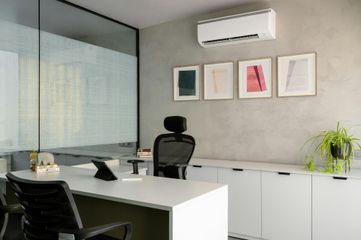 A modern office with a desk, two ergonomic chairs, wall art, a potted plant, and a wall-mounted air conditioner.