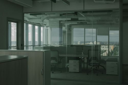 A modern office interior with glass-walled cubicles, desks, and chairs overlooking a city skyline.