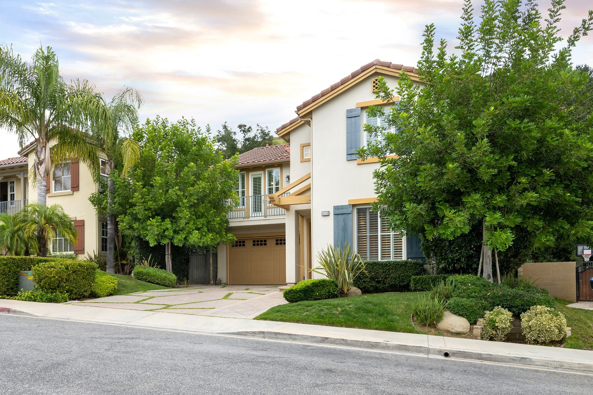 Two-story home with light tan stucco, blue shutters, and a beige garage door. Trees and green bushes surround it.