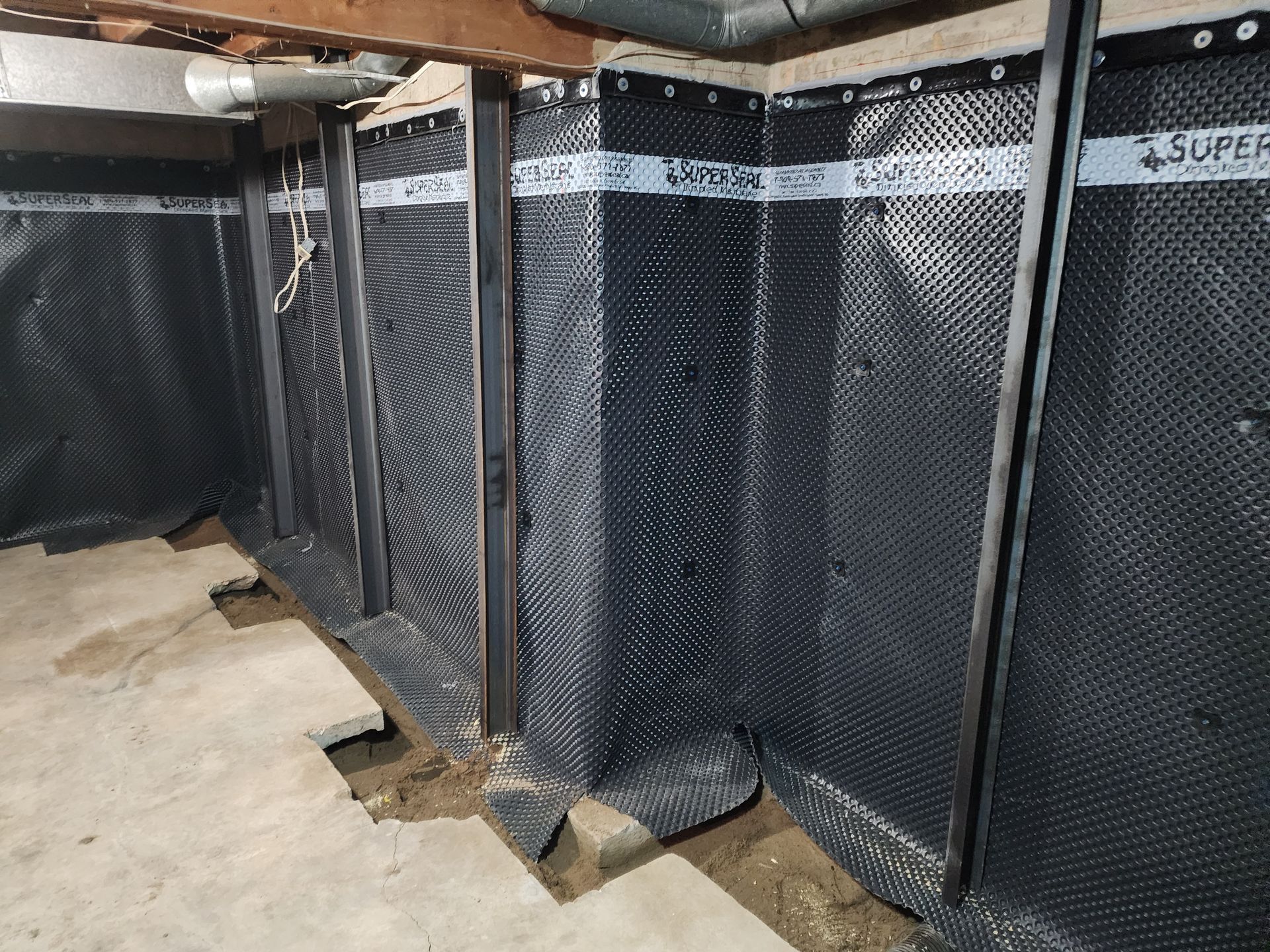Basement interior with black dimpled drainage membrane on walls, support beams, and exposed concrete floor.