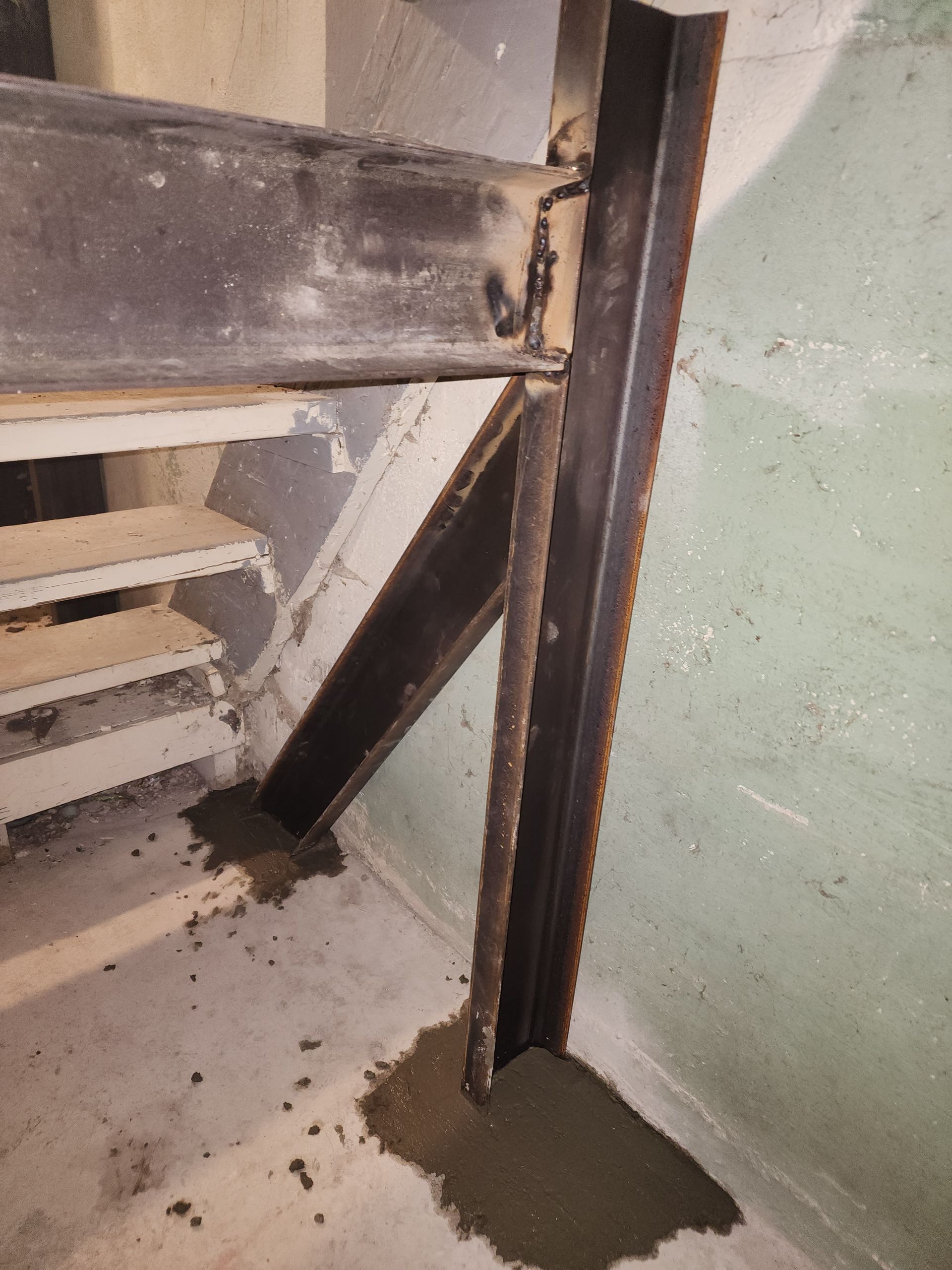 Steel beams supporting a staircase. A vertical beam is attached to an angled beam and floor, with concrete around the base.