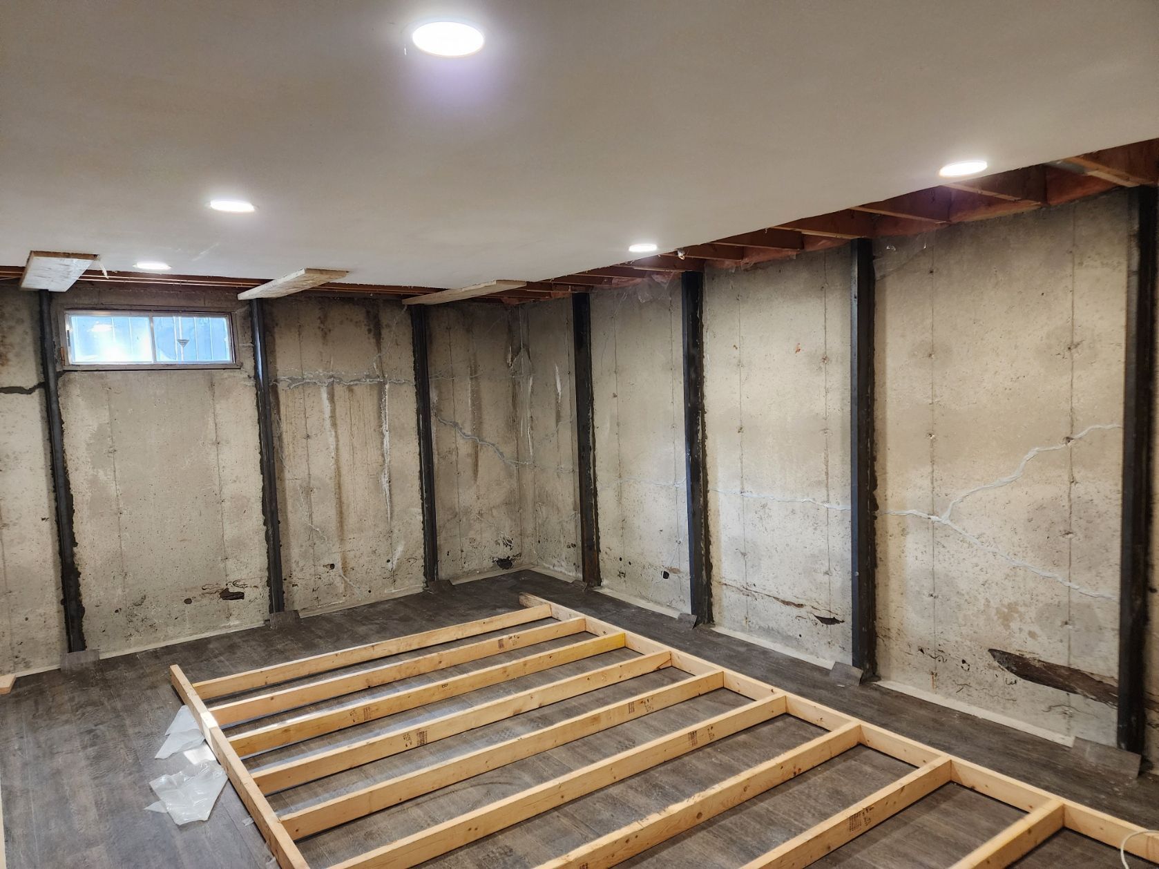 Basement room under construction: concrete walls, wood framing on floor, and exposed ceiling beams.