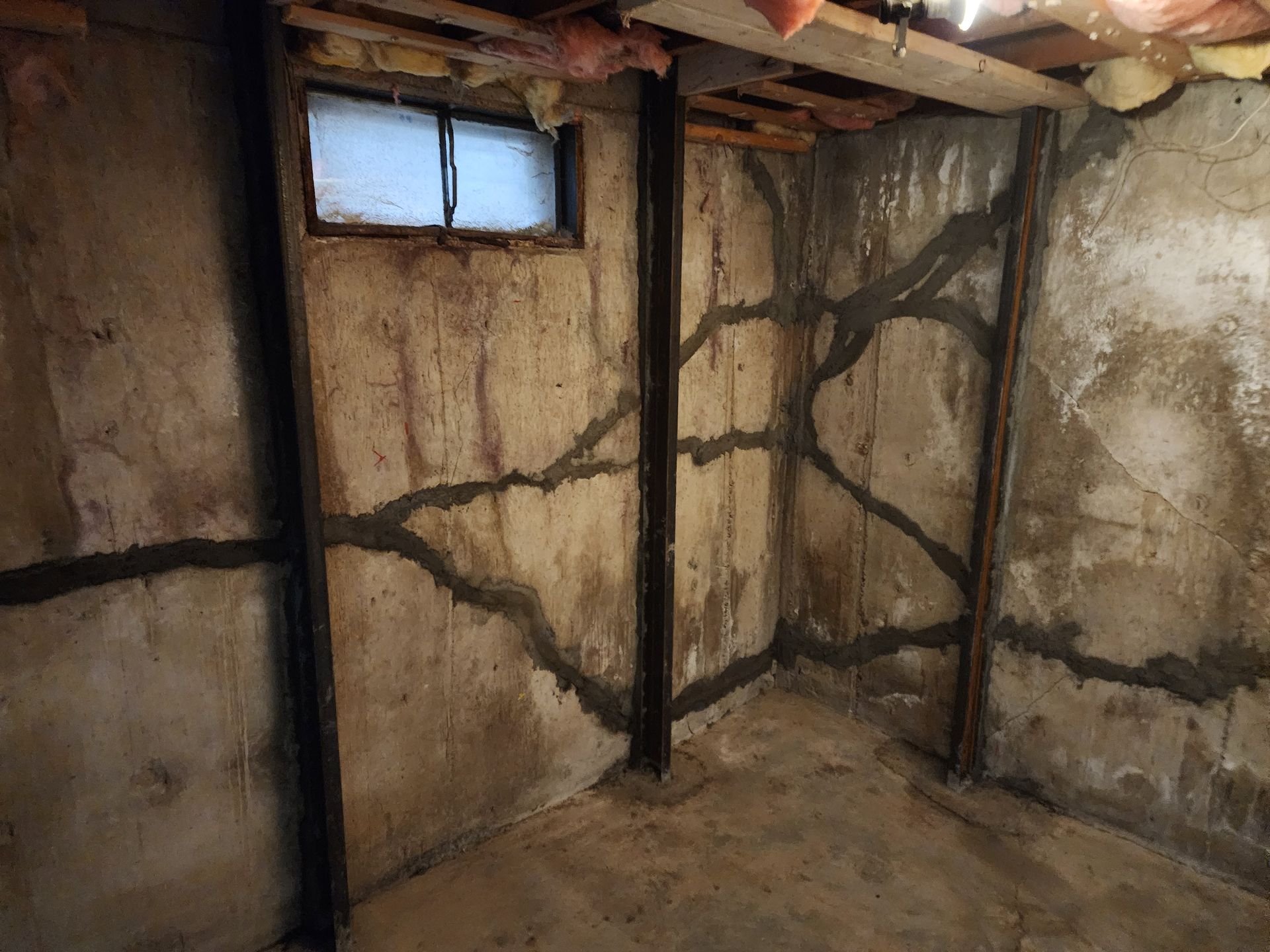 Basement interior with repaired foundation cracks. Concrete walls, metal beams, small window, and grey crack sealant.