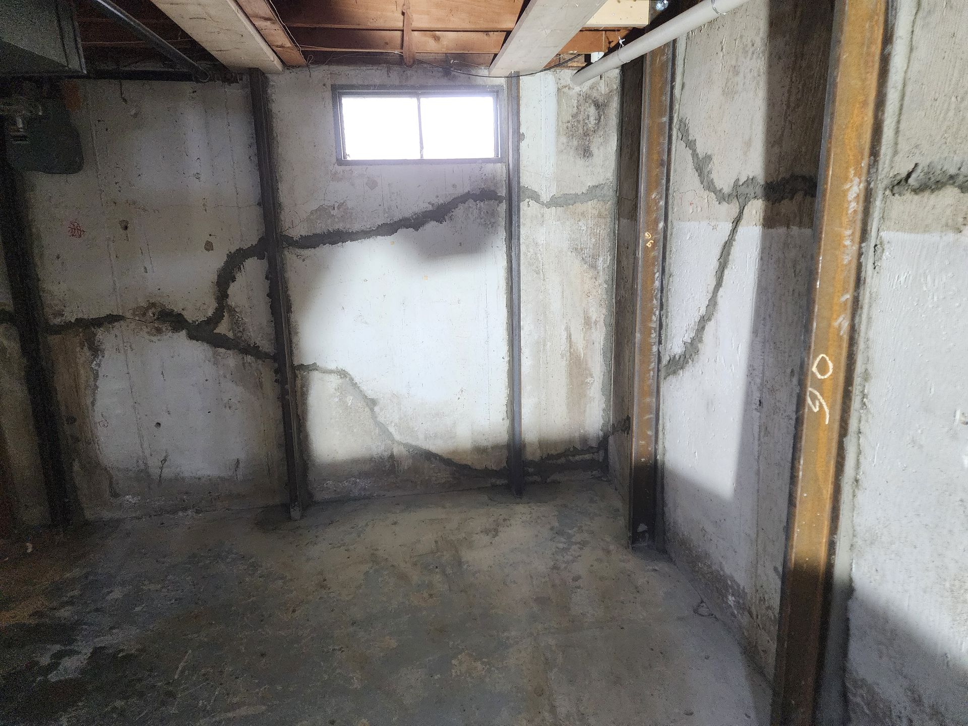 Cracked basement wall with steel supports, window above. Grey concrete, water stains.