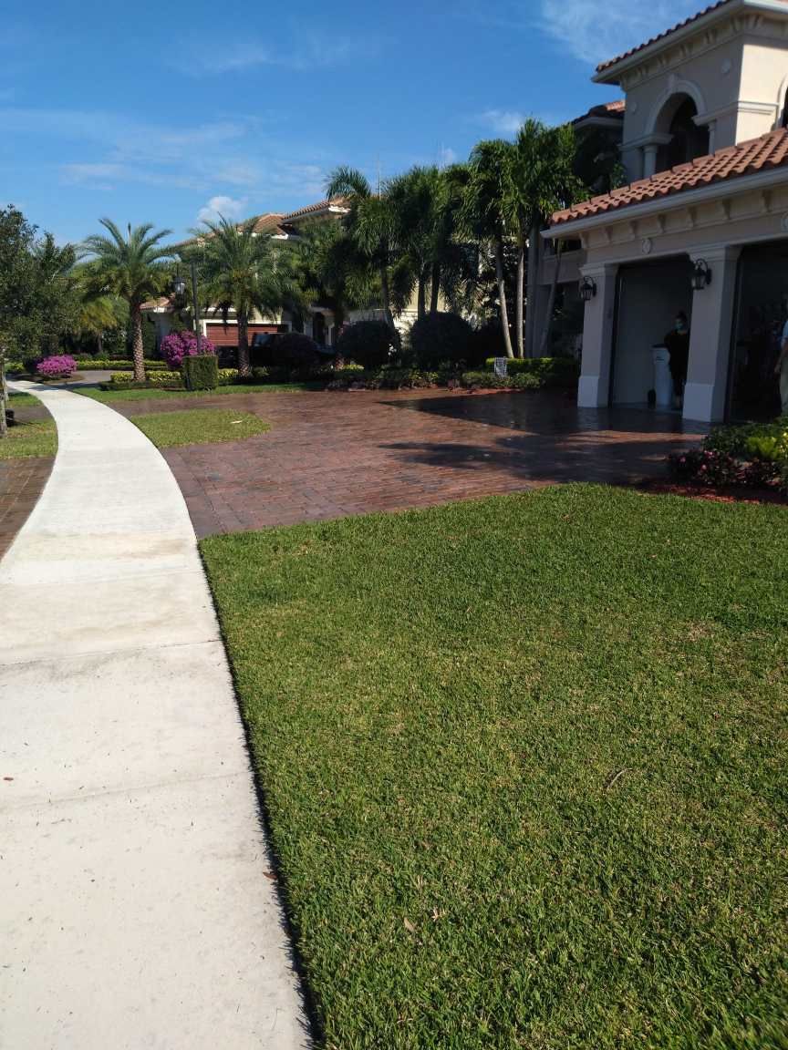 A concrete walkway leading to a large house with a lush green lawn.