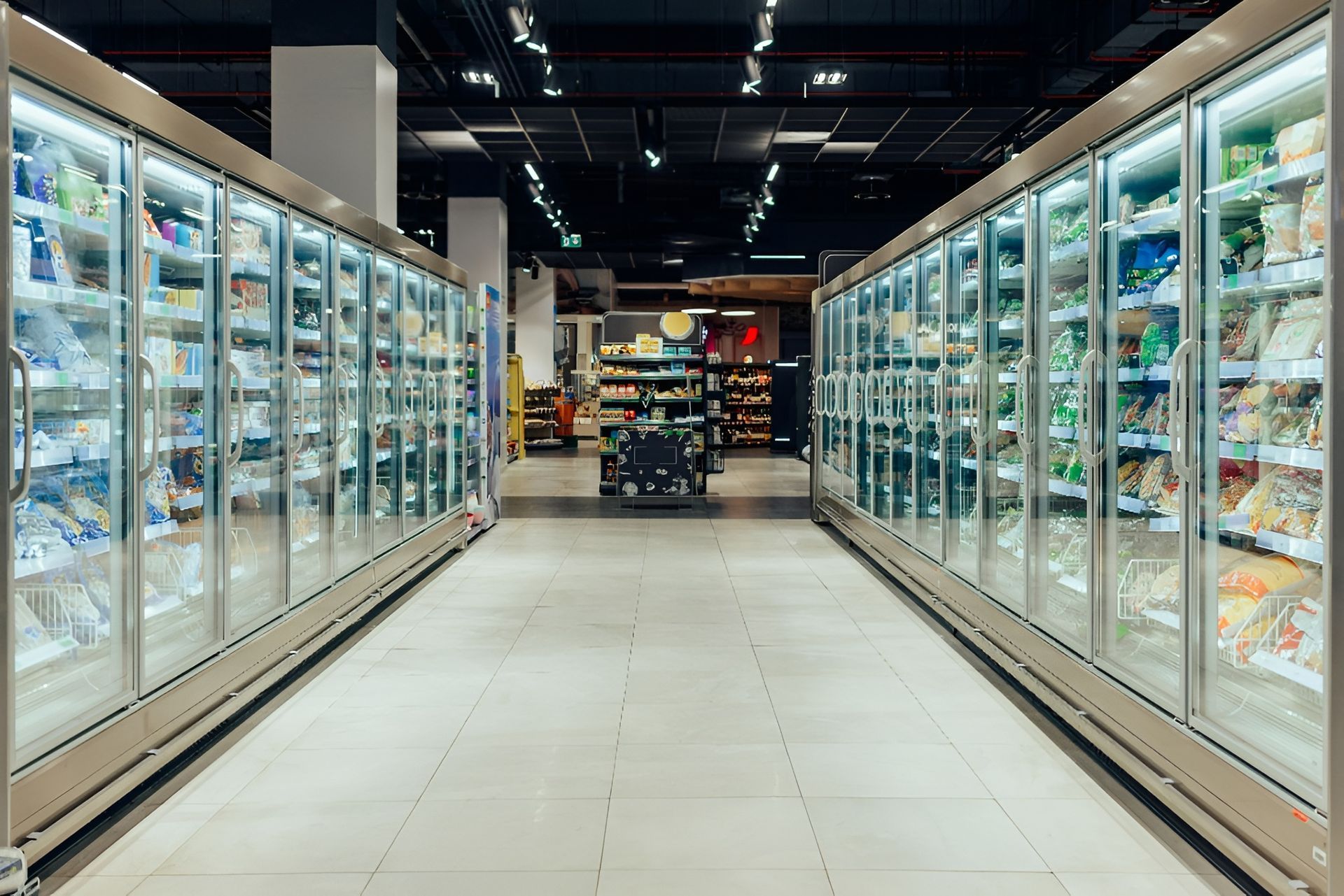 Rows of glass-door freezers in a supermarket aisle filled with frozen food.