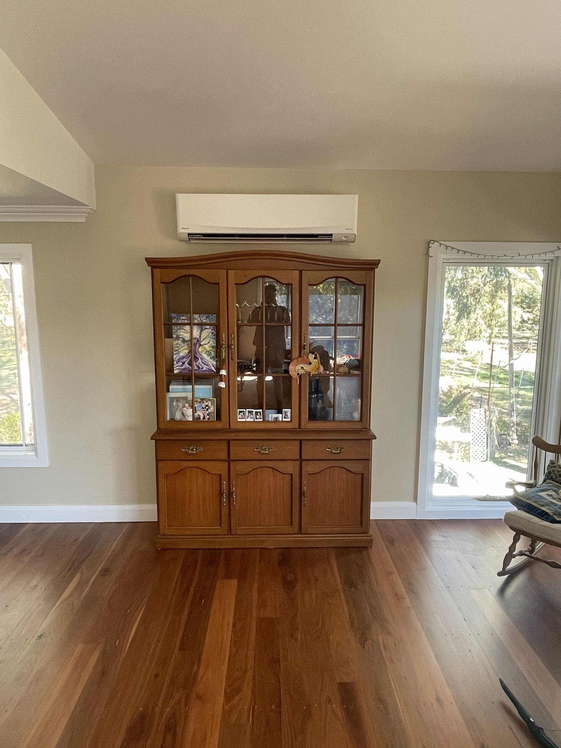 A wooden hutch with glass doors sits below a white air conditioning unit, next to a window.