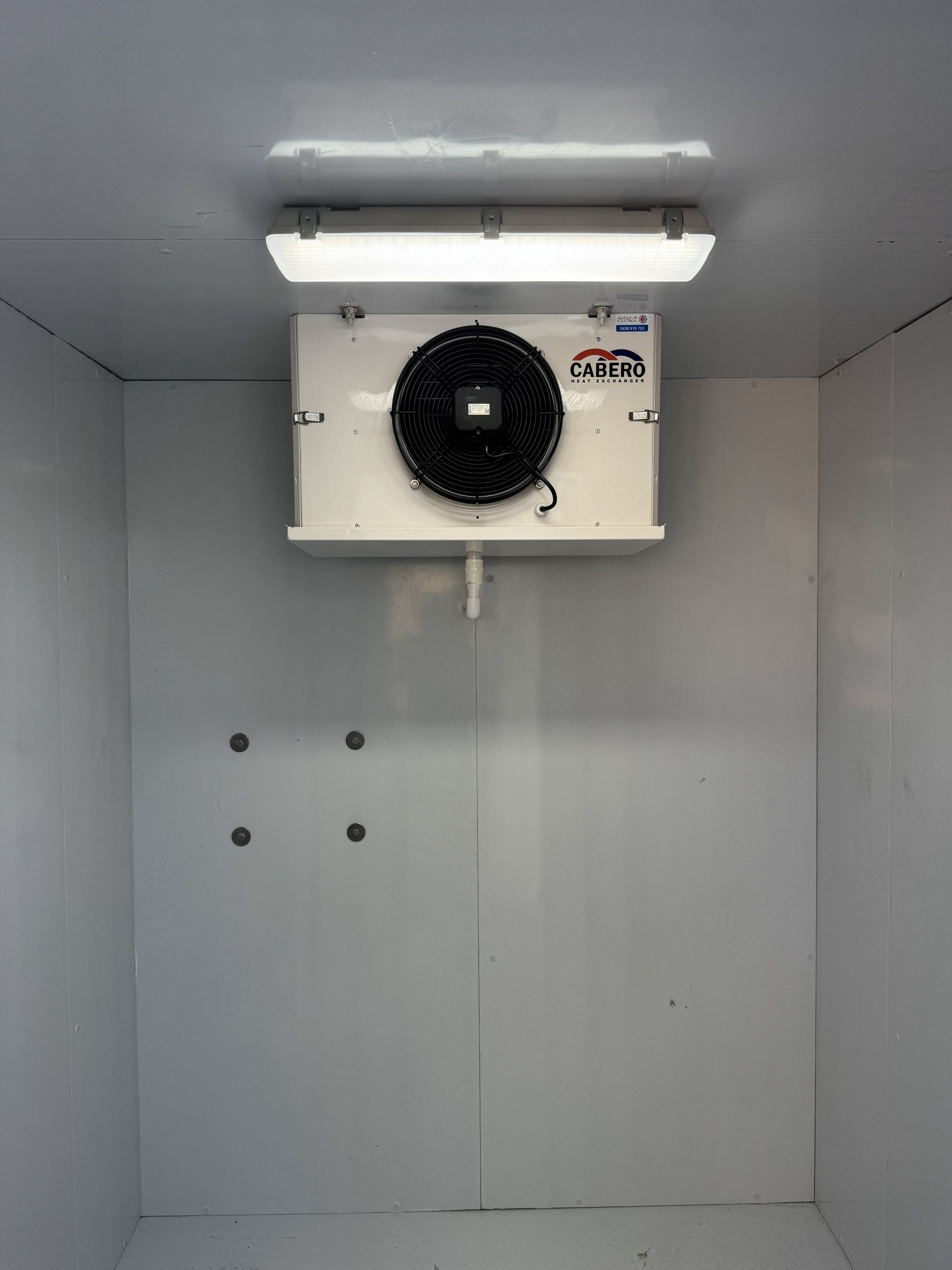 Inside a white walk-in cooler, a cooling unit with a black fan is mounted on the wall.