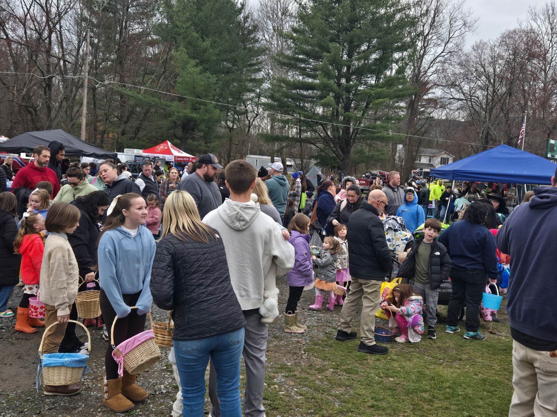 Crowd at an outdoor Easter egg hunt. Children and adults with baskets on a grassy area near trees and tents.