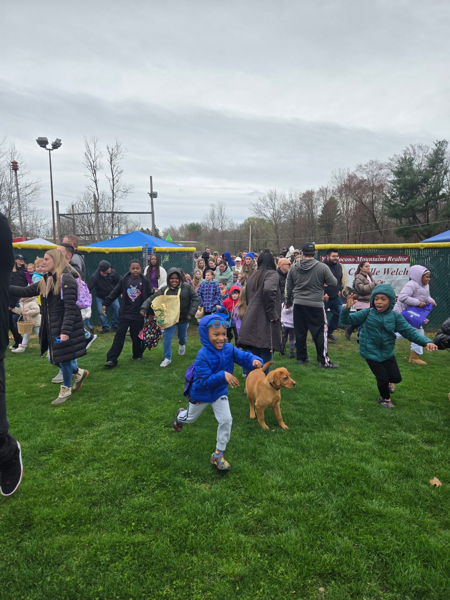 Children running on grass, collecting items, possibly an Easter egg hunt. Adults watch.