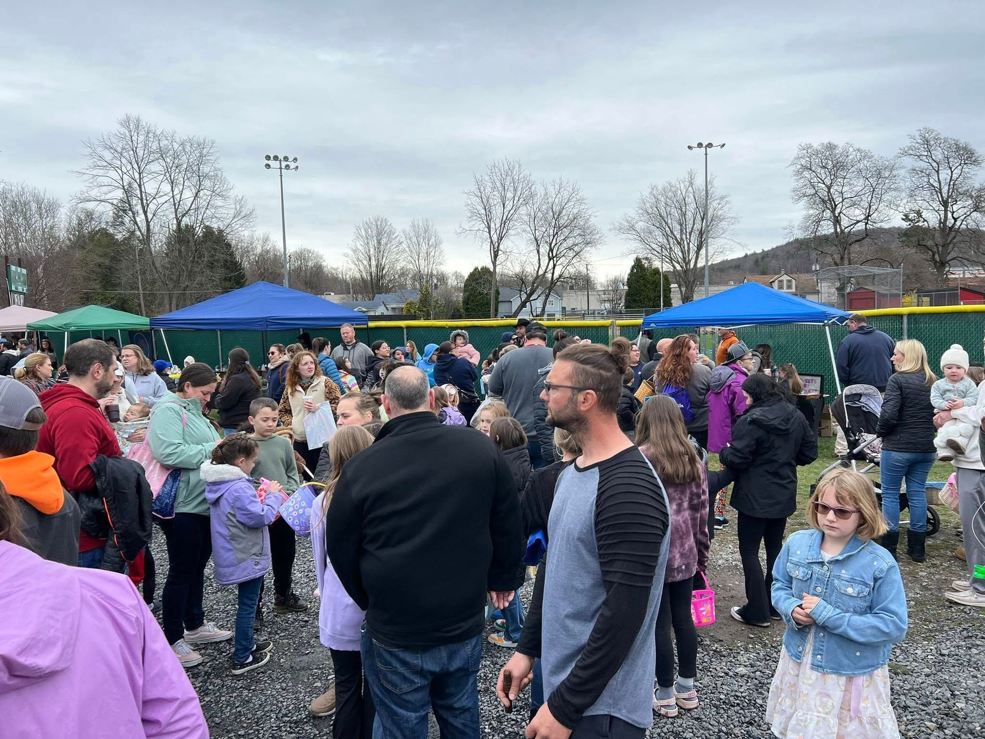 Crowd of people gathered outdoors under cloudy sky; blue and green tents in background.
