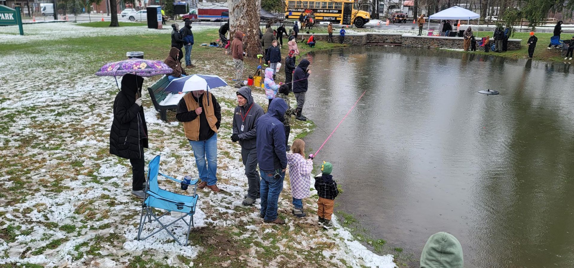 People fishing at a pond in a park on a snowy day, some with umbrellas.