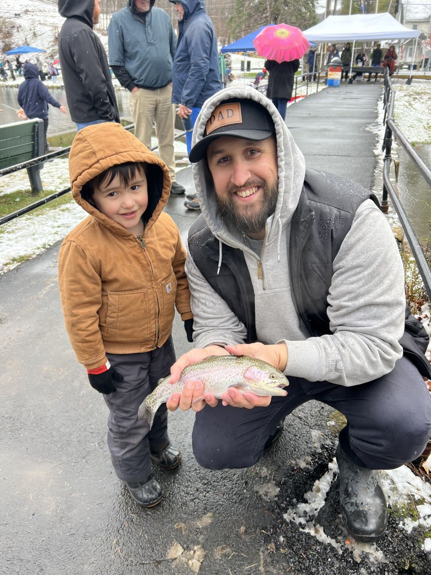 Man and child pose with a fish outdoors. Man kneels, holding fish. Child stands, smiles. Snowy ground.