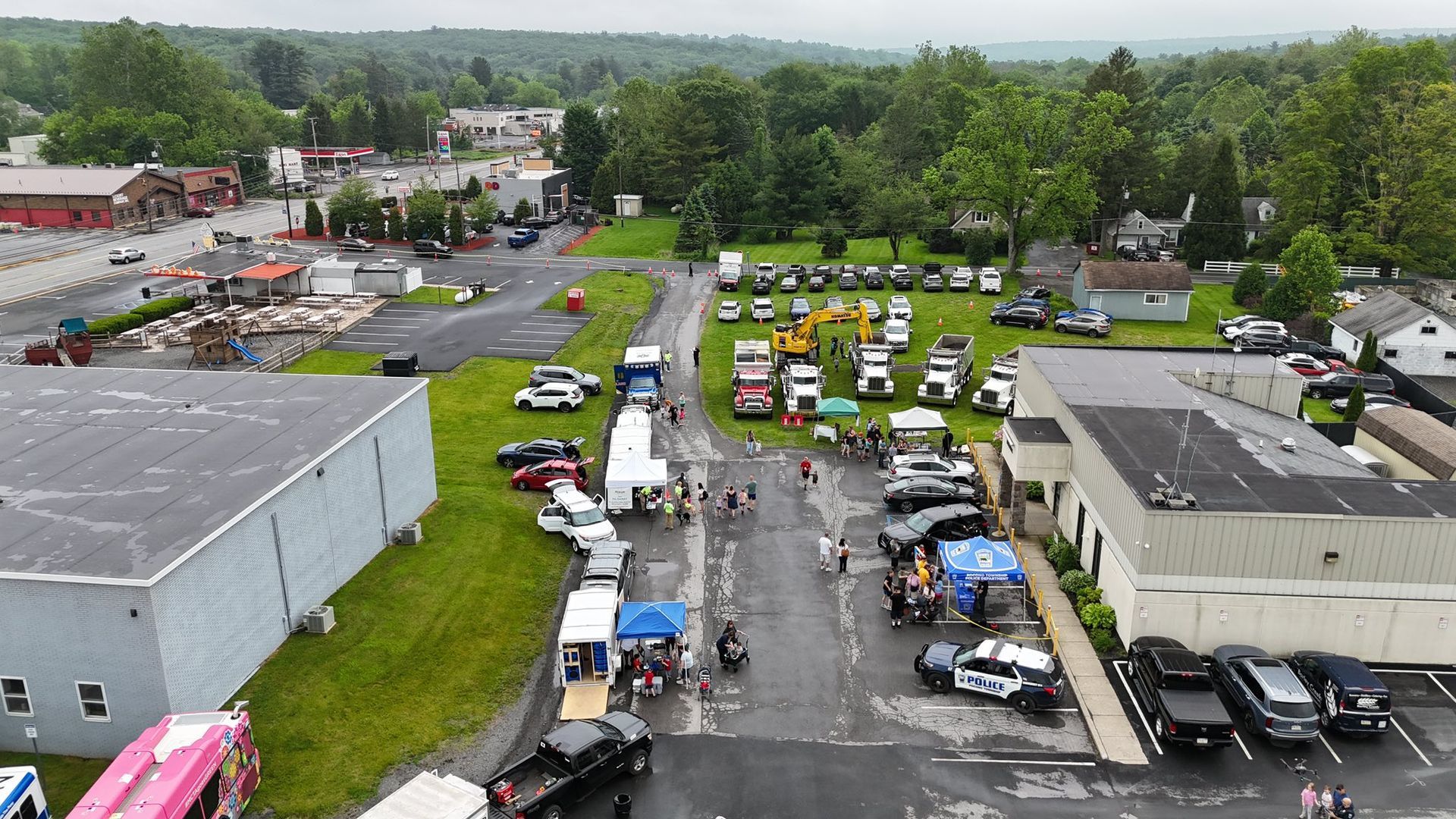 Aerial view of outdoor event: vehicles, tents, people gathering in a town, cloudy sky.