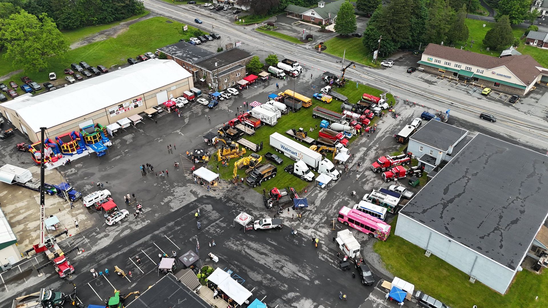 Aerial view of an event with parked vehicles, vendor stalls, and buildings on a paved area.