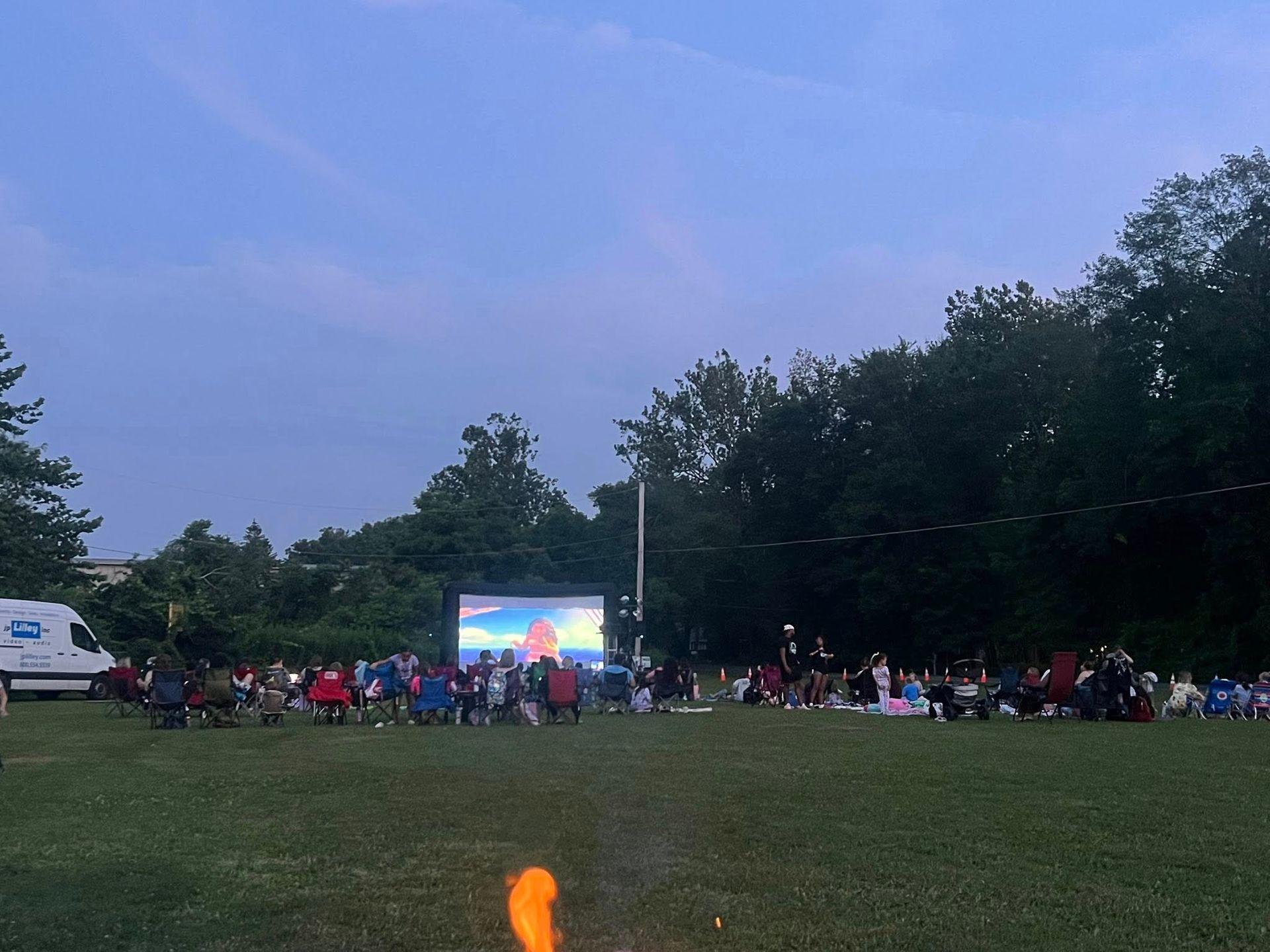 People watching a movie outdoors on a large screen in a grassy field at dusk.