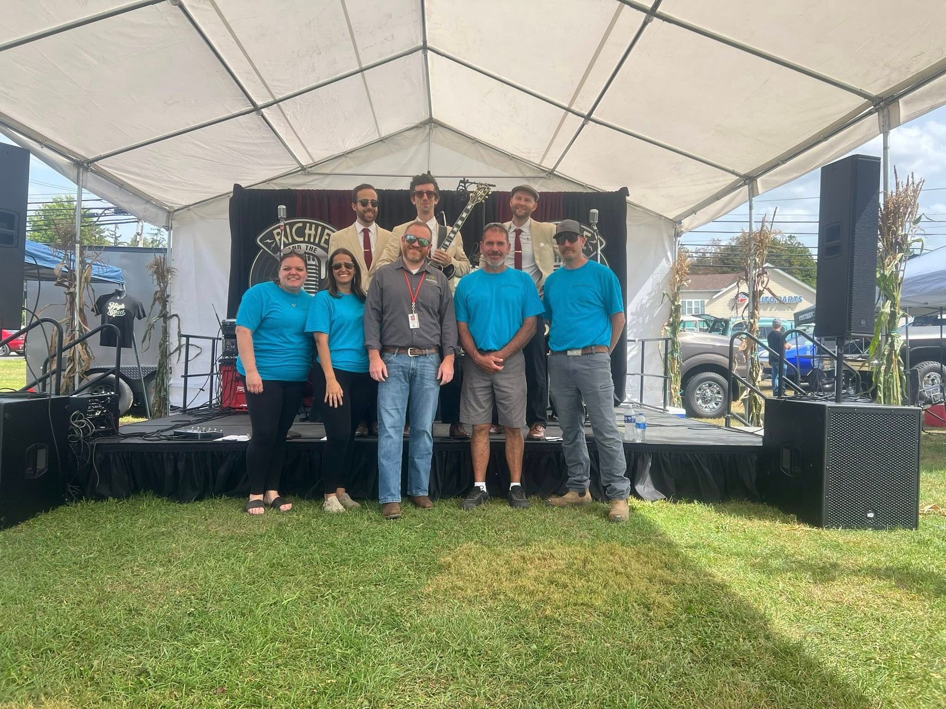 Band and crew pose on stage under a white tent in an outdoor setting.