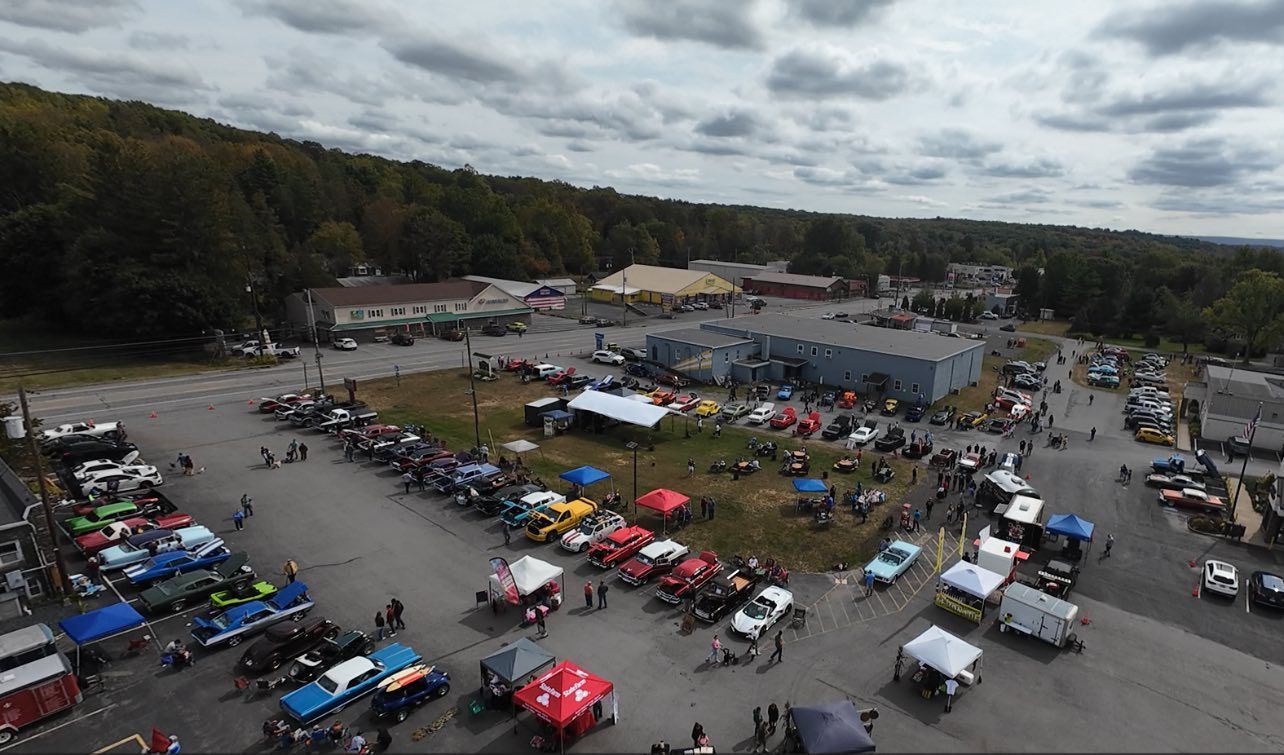 Car show outdoors; many vehicles, tents, people in parking area. Forest in background, cloudy sky.