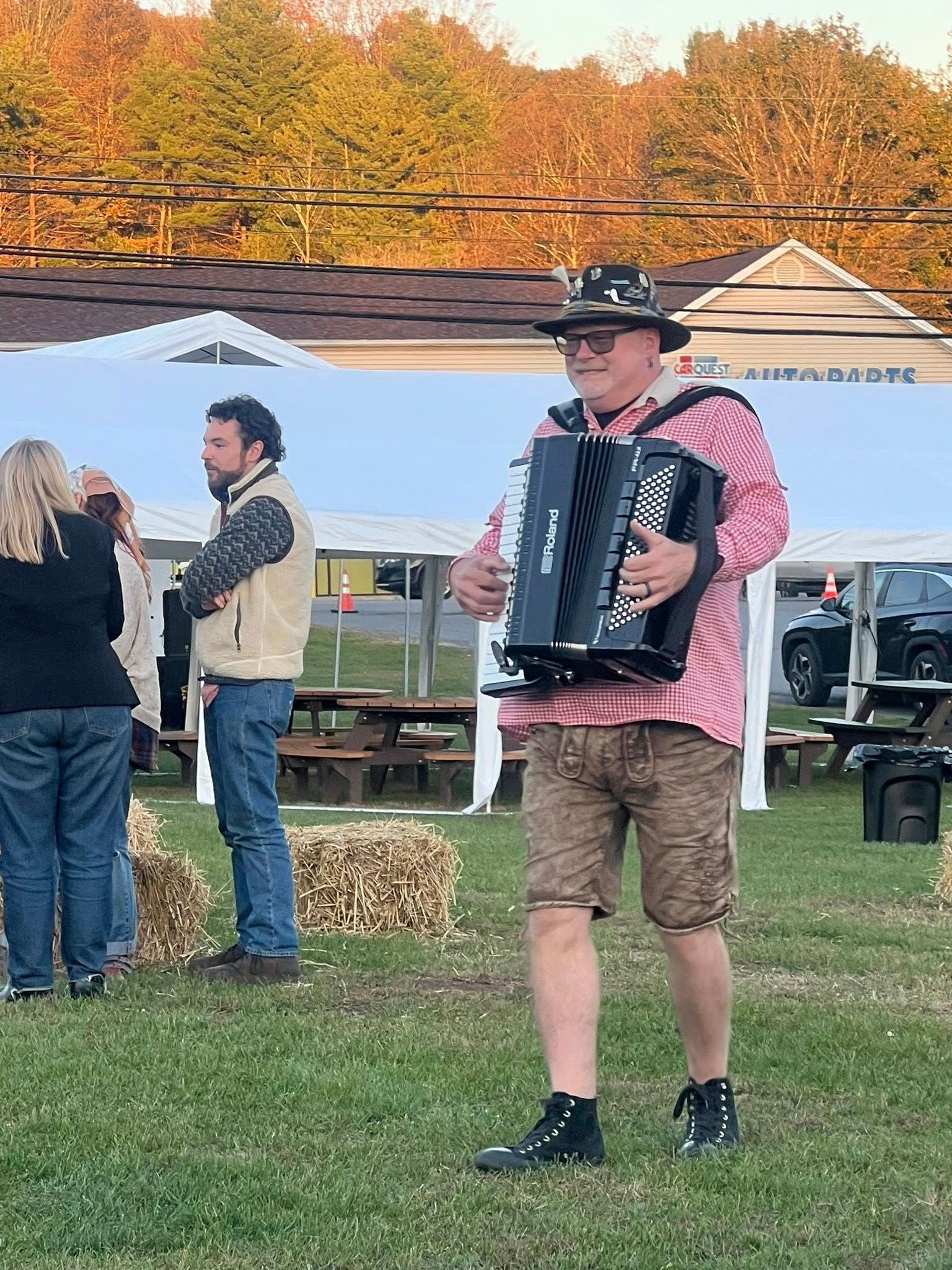 Man in lederhosen plays accordion outside. People mill about. A white tent is in the background.