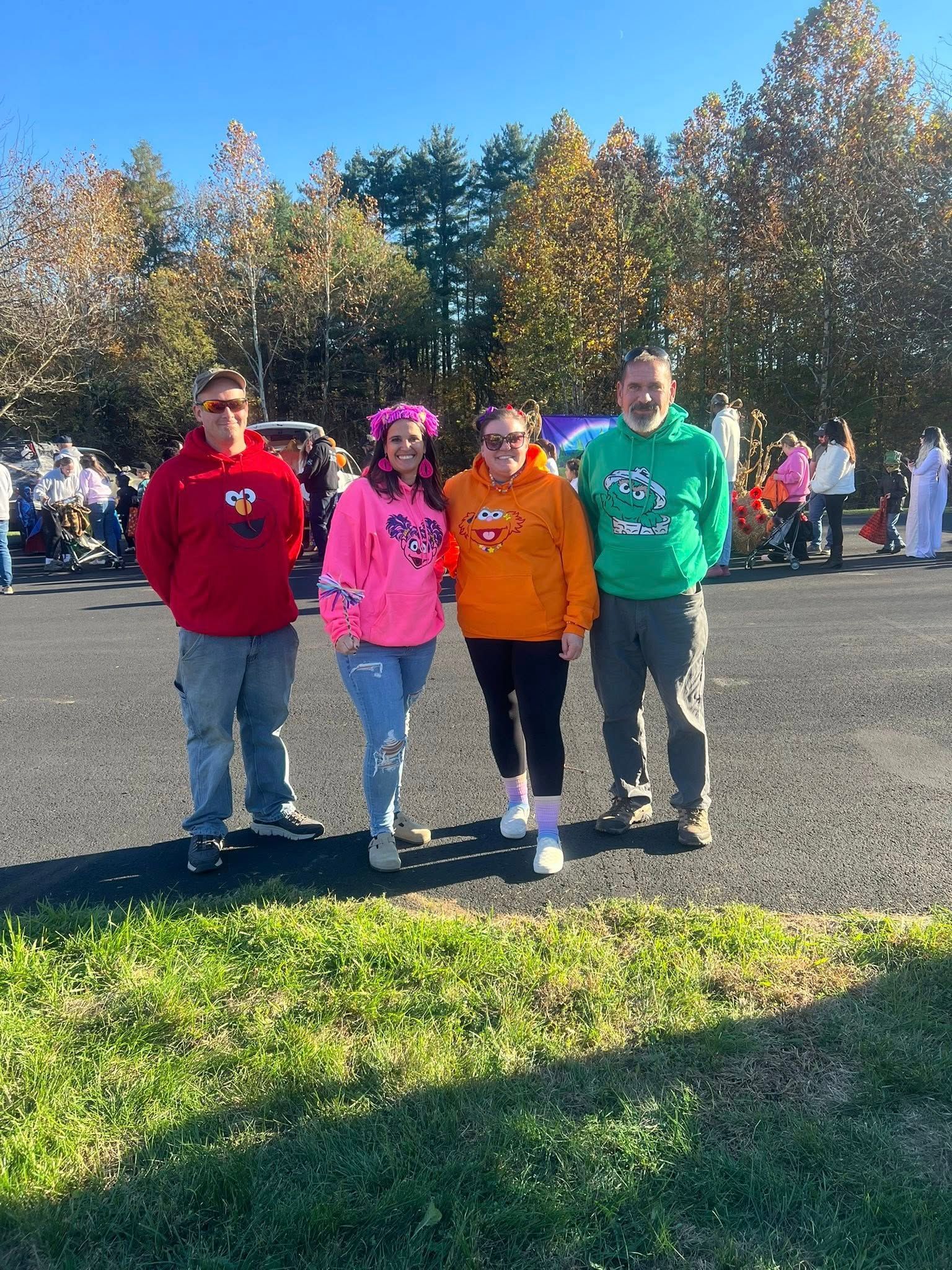 Four people in hoodies standing outdoors. One pink, one orange, one red, and one green. Trees and a crowd in the background.