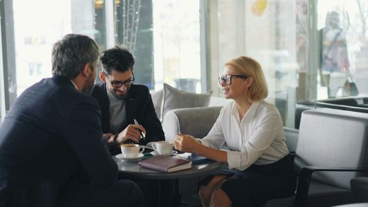 A man and a woman are sitting at a table talking to a man in a suit.