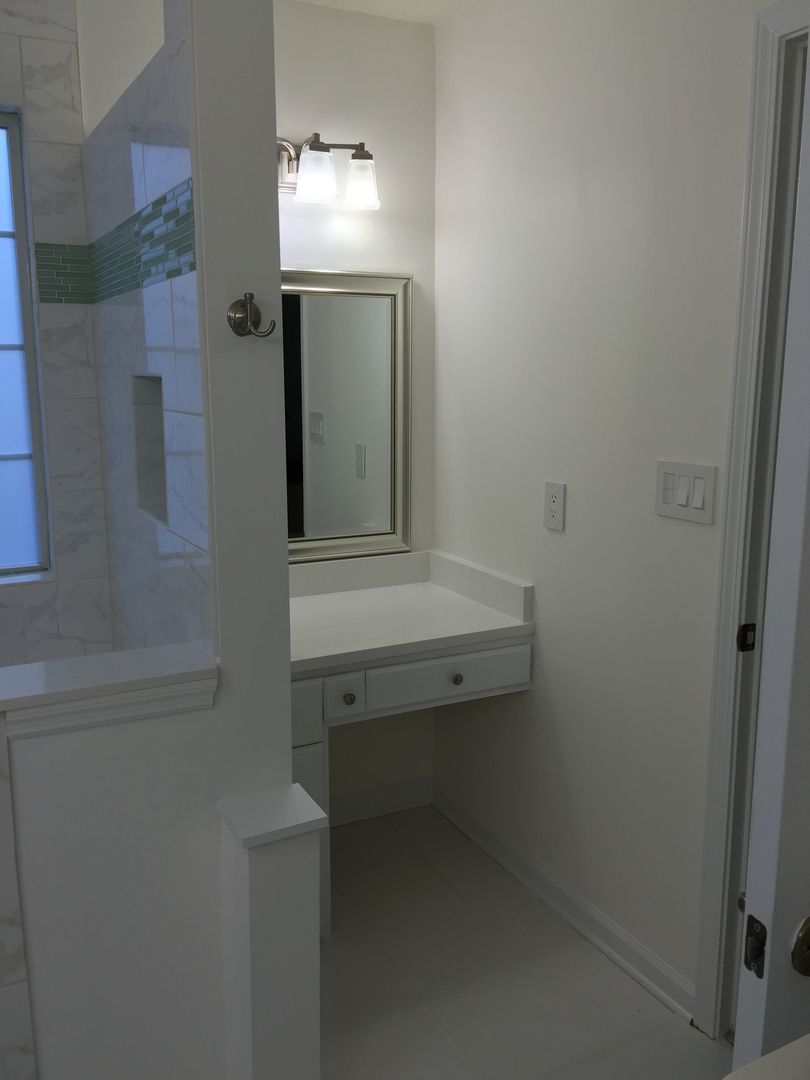 Small white bathroom vanity with mirror and light fixture. Adjacent shower on left. Door on right.