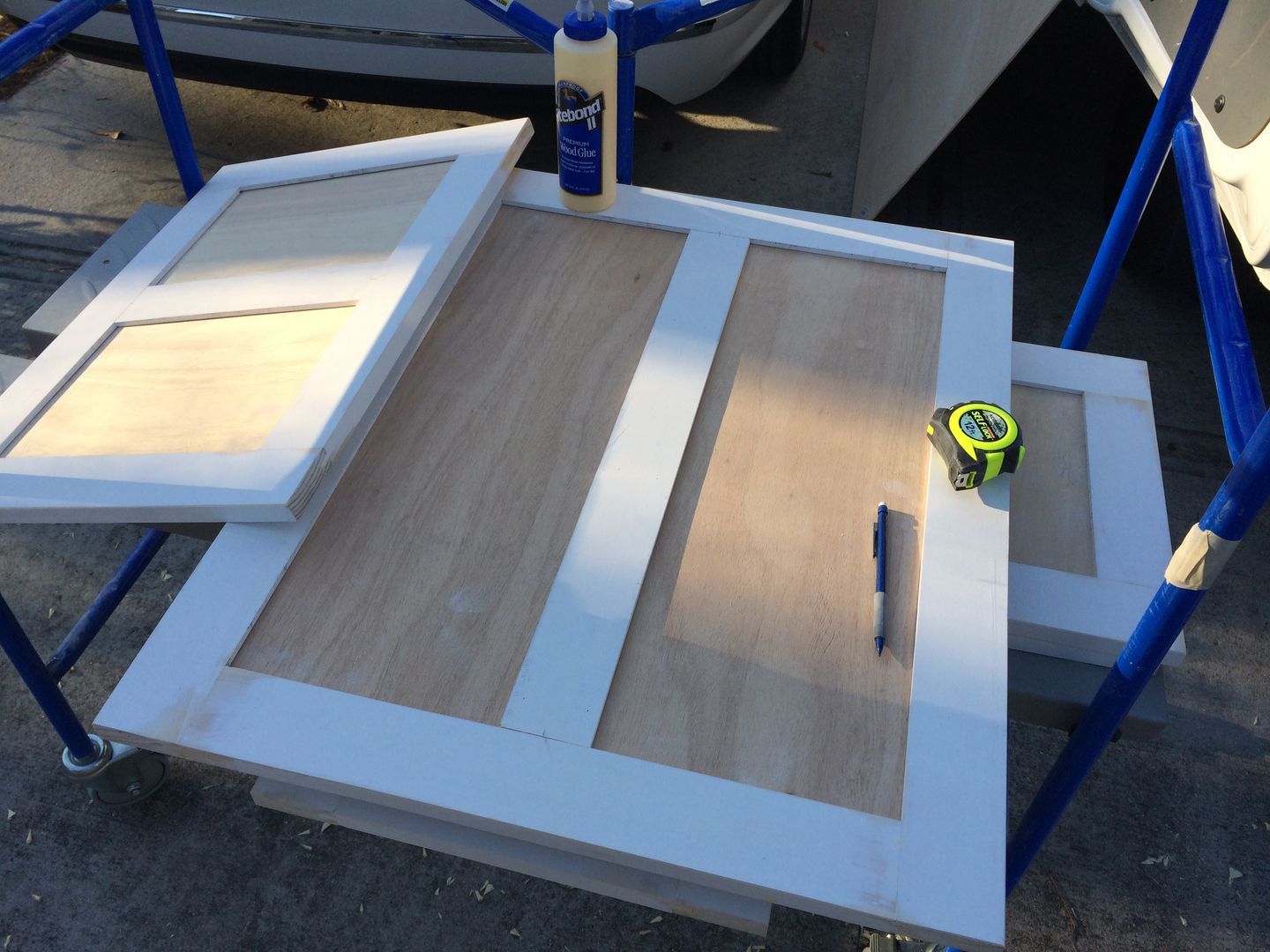 Cabinet door construction: white frames on light wood panels, with glue, tape measure, and pen on blue cart.
