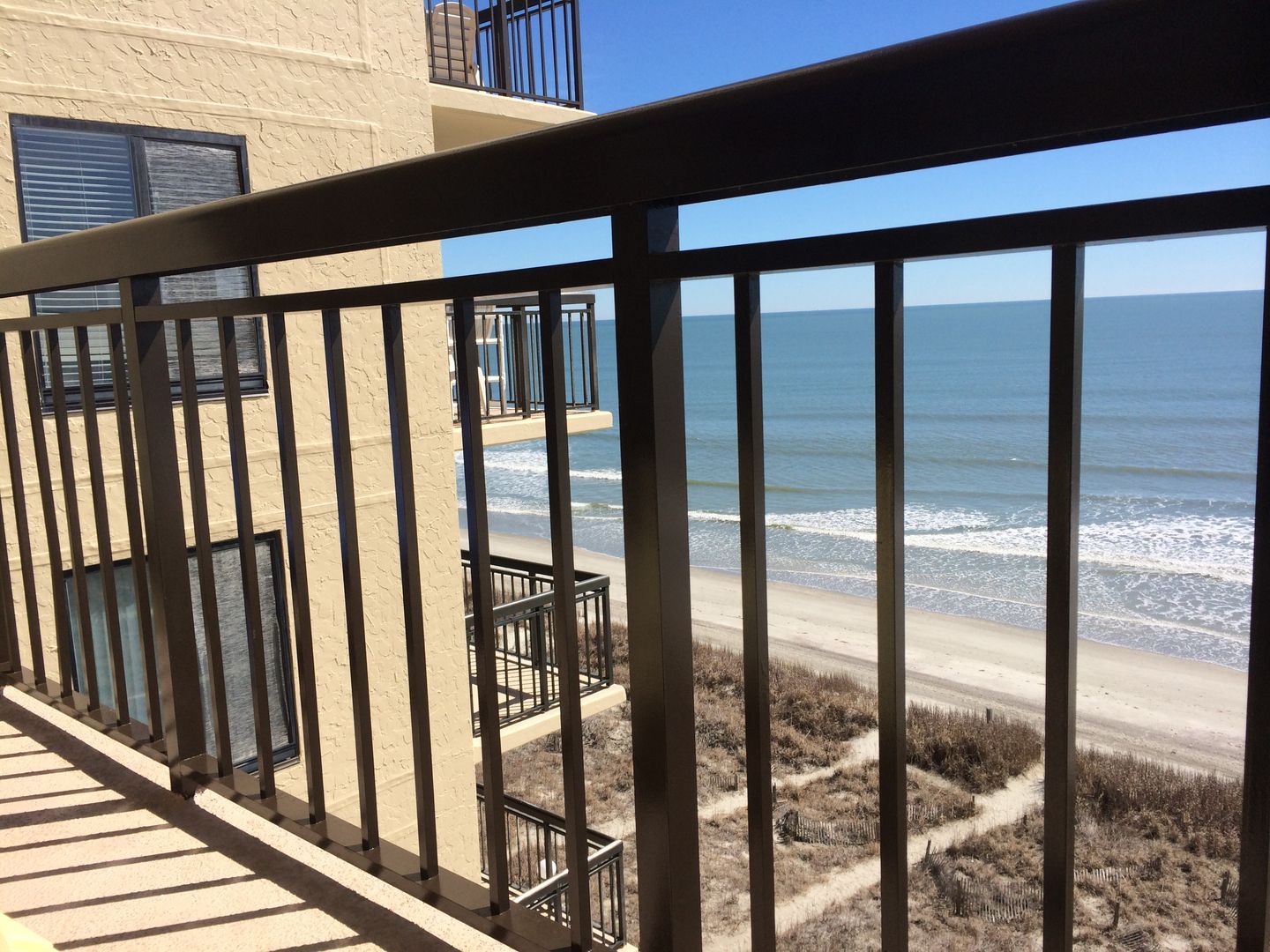 Ocean view from a balcony with black metal railing. Sandy beach and blue sky visible.