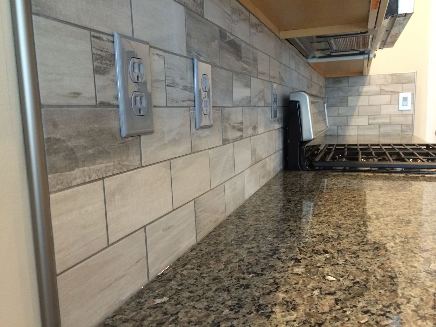 Kitchen backsplash with tan and grey tiles, electrical outlets, and granite countertop.