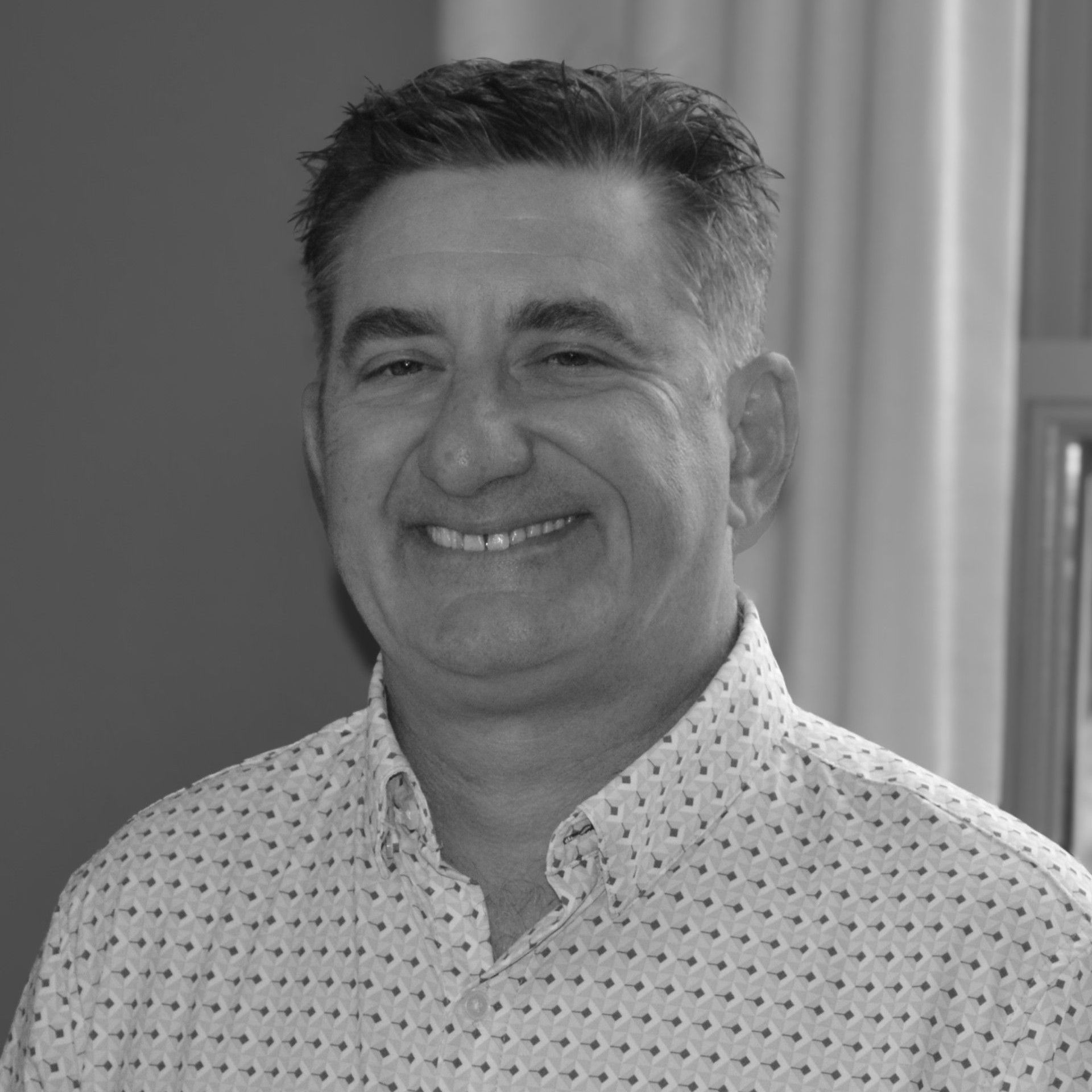 A man wearing a gray sweater and a blue shirt is smiling in front of a brick wall.