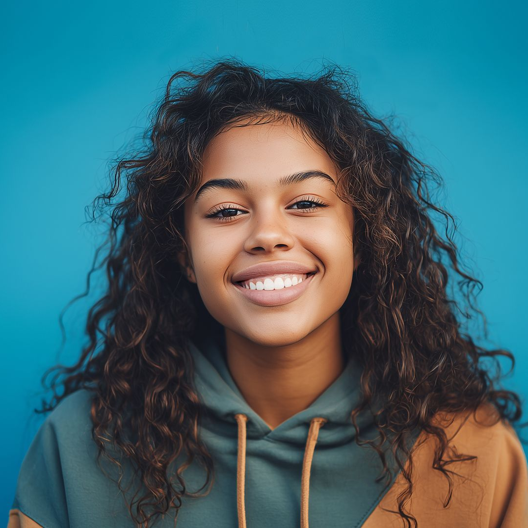 Young Black woman with curly hair smiles, wearing a green and brown hoodie against a blue background.