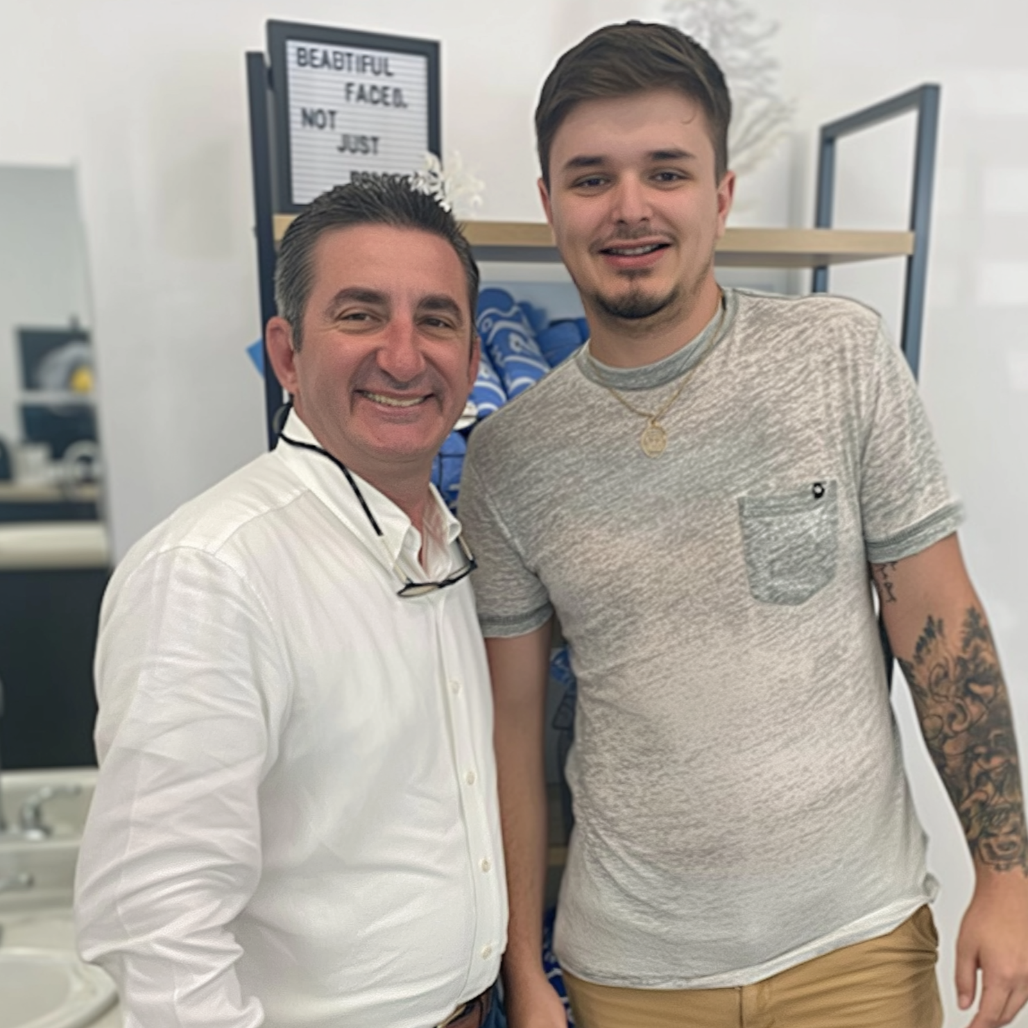 Two men smiling, standing near a shelf with towels. One man wears a white shirt, the other a gray shirt.