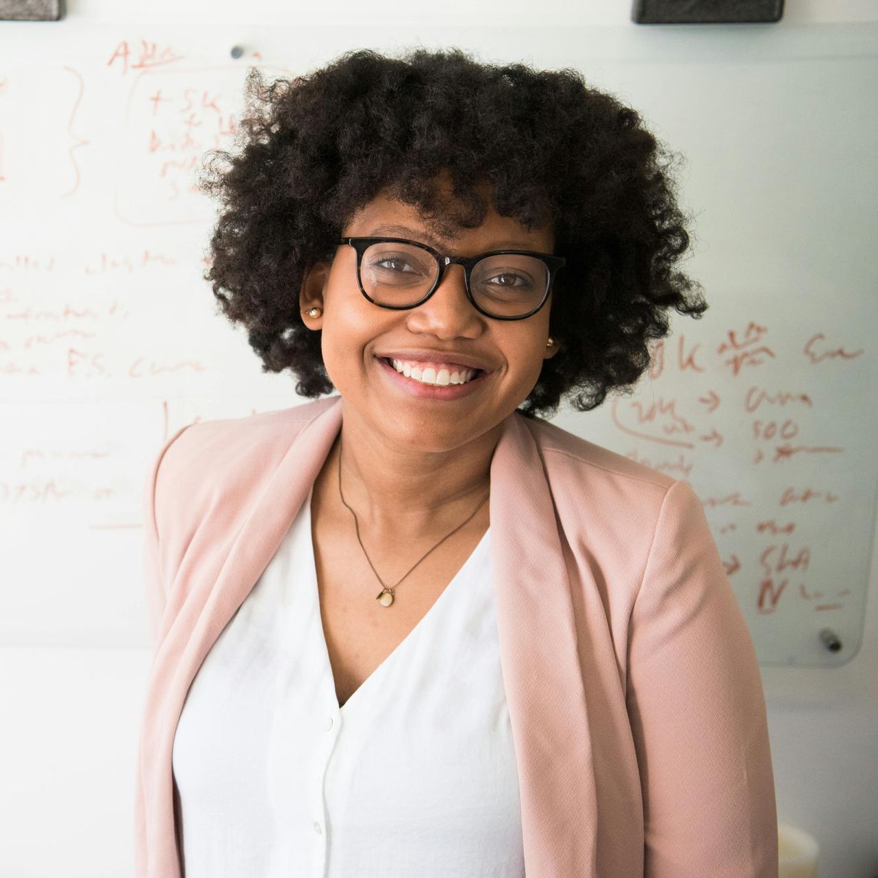 A woman wearing glasses and a pink jacket smiles in front of a white board