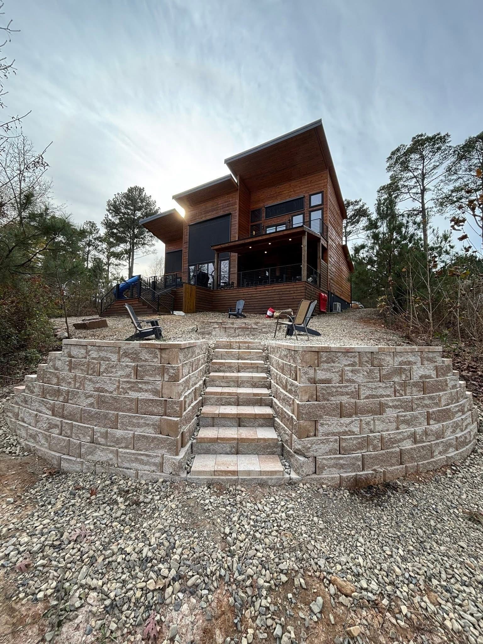 A modern wooden cabin sits atop a stone retaining wall with a central staircase, surrounded by woods under a sunny sky.