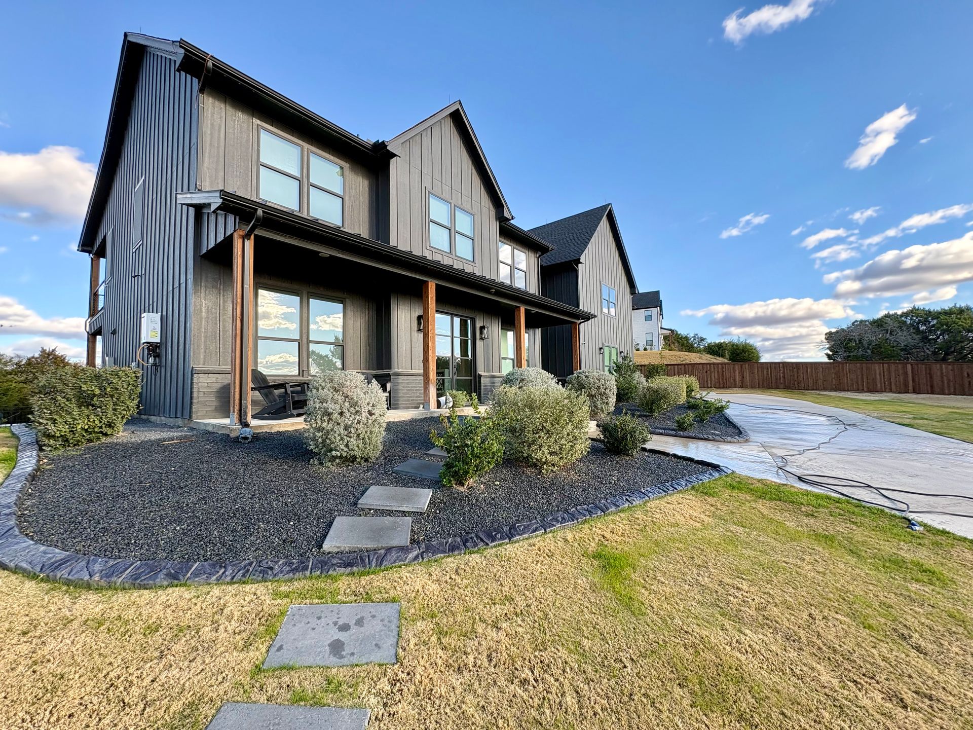 A modern, dark-wood two-story house with a covered porch, stone walkway, and dark mulch landscaping under a blue sky.