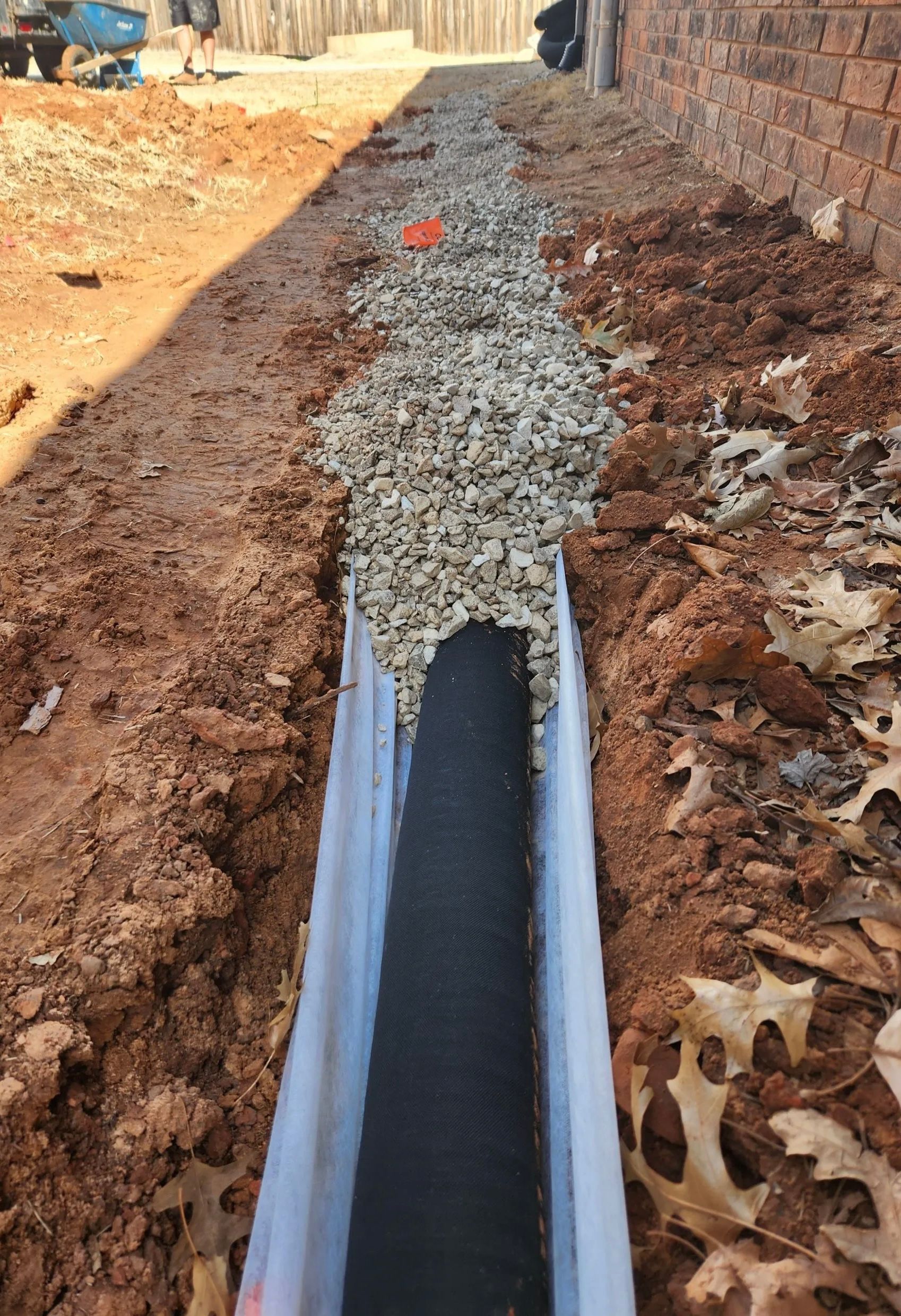 A drainage pipe wrapped in black filter fabric sits inside a trench filled with gravel along a brick house foundation.