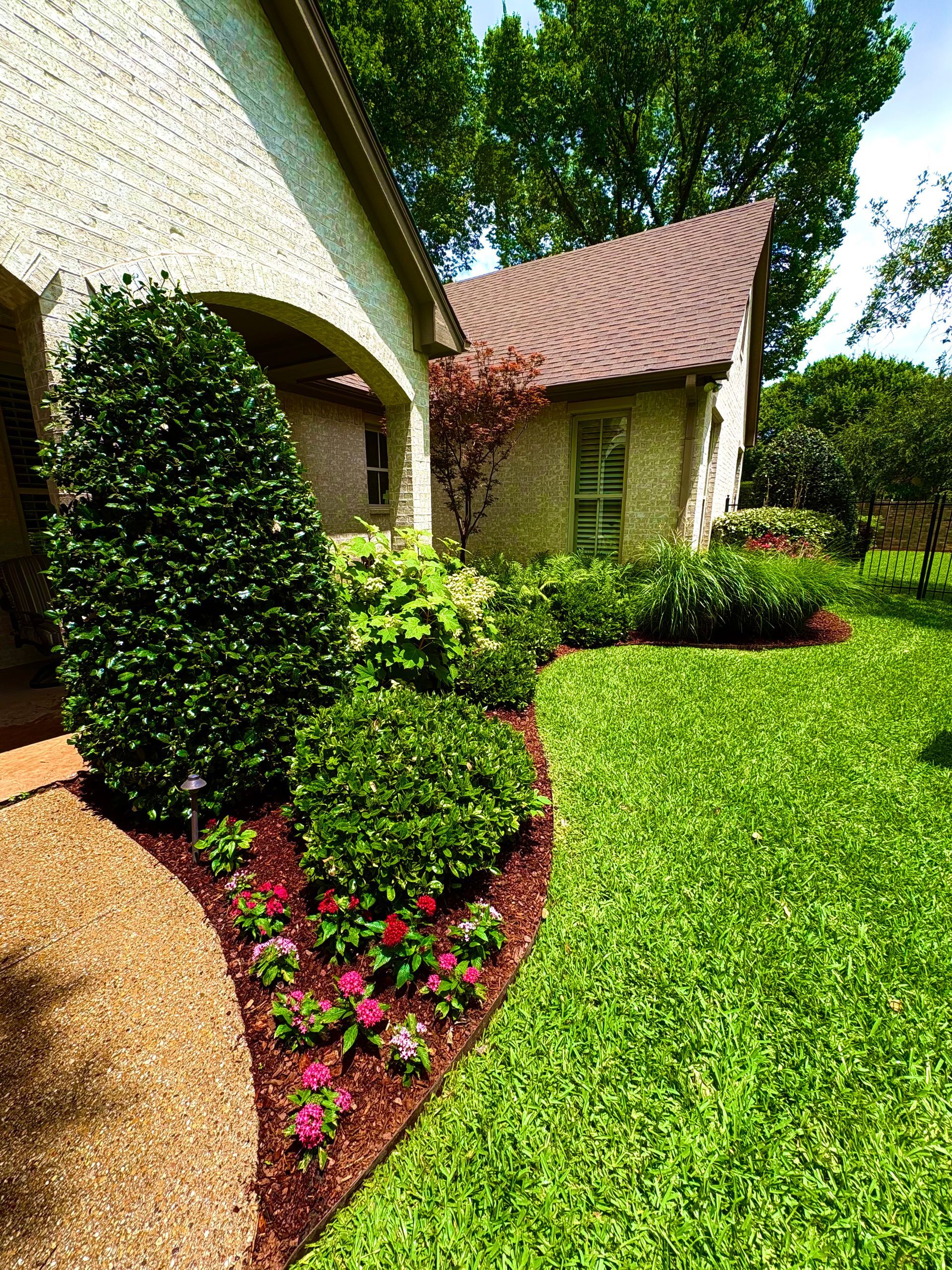 A stone house with a landscaped garden bed containing shrubs and small pink flowers, adjacent to a manicured lawn.