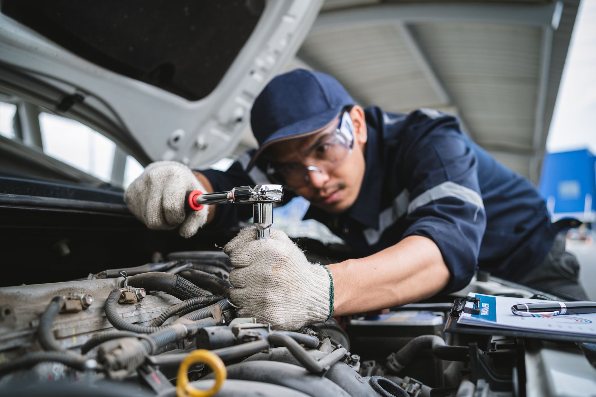 A male auto repair shop worker uses a tool to repair and inspect a car with its hood up.