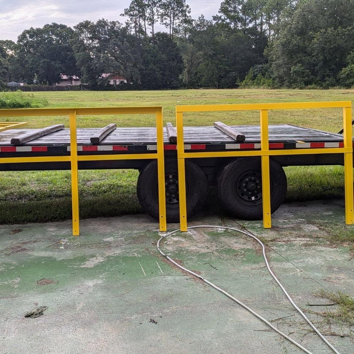 Yellow handrails on a black trailer, parked on grass. Trees and a building in the background.
