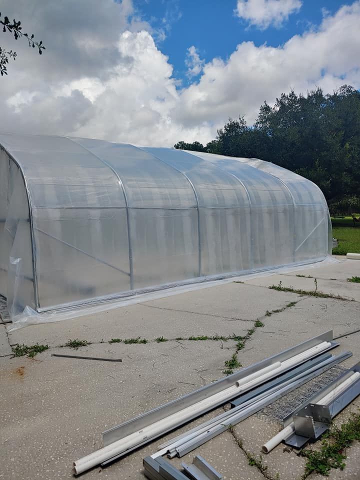 A large, arched greenhouse made of clear plastic, on a concrete pad. Partially assembled metal pieces are nearby.