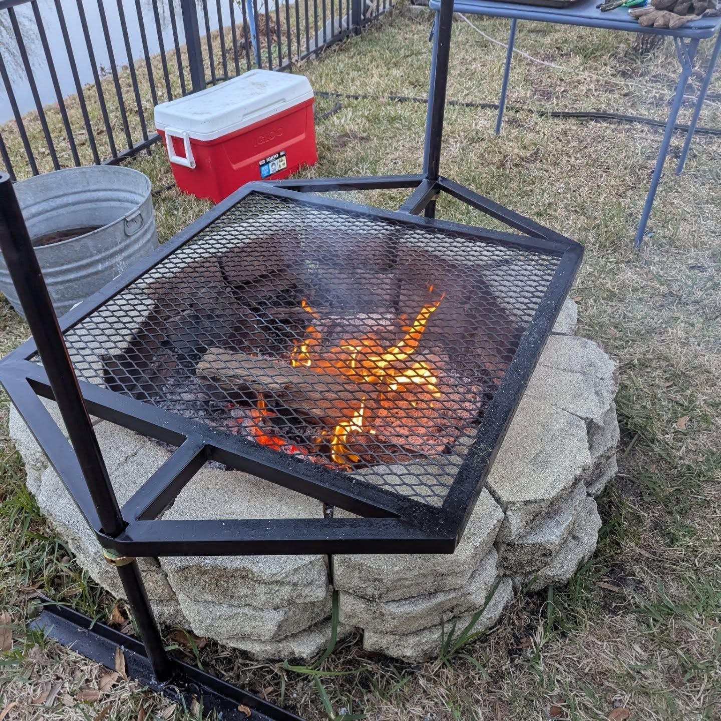 Fire pit with metal grate and burning logs, surrounded by stone blocks, outdoors.