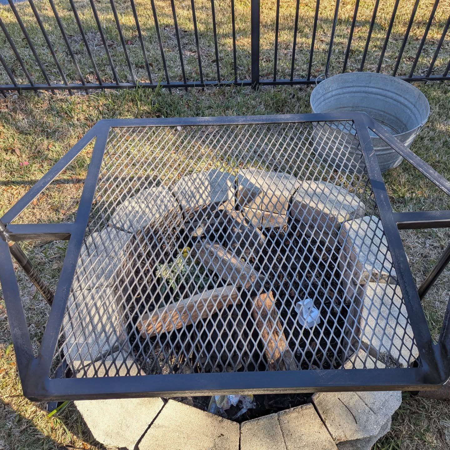 Fire pit with metal grate and rocks, surrounded by a metal fence and grass.