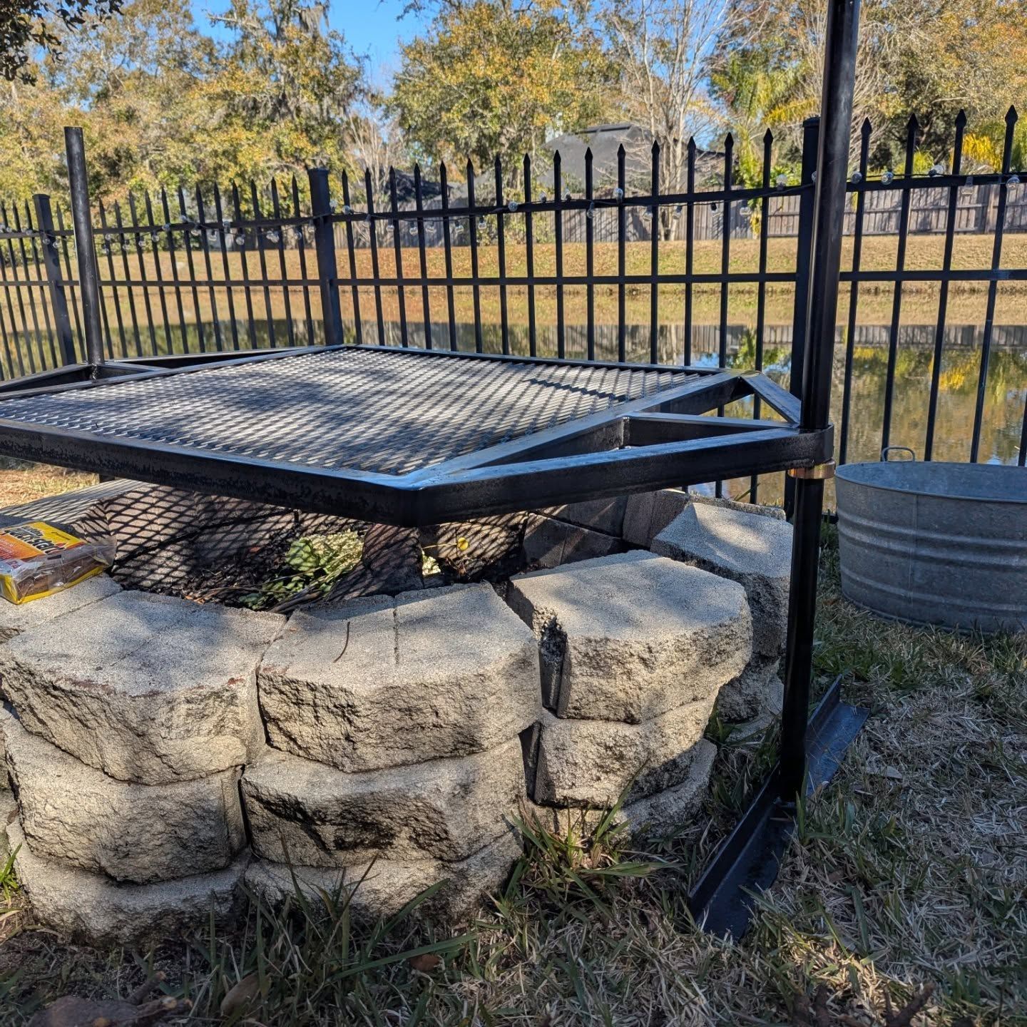 Metal grilling structure over a stone fire pit, with a metal bucket and black fence in the background.