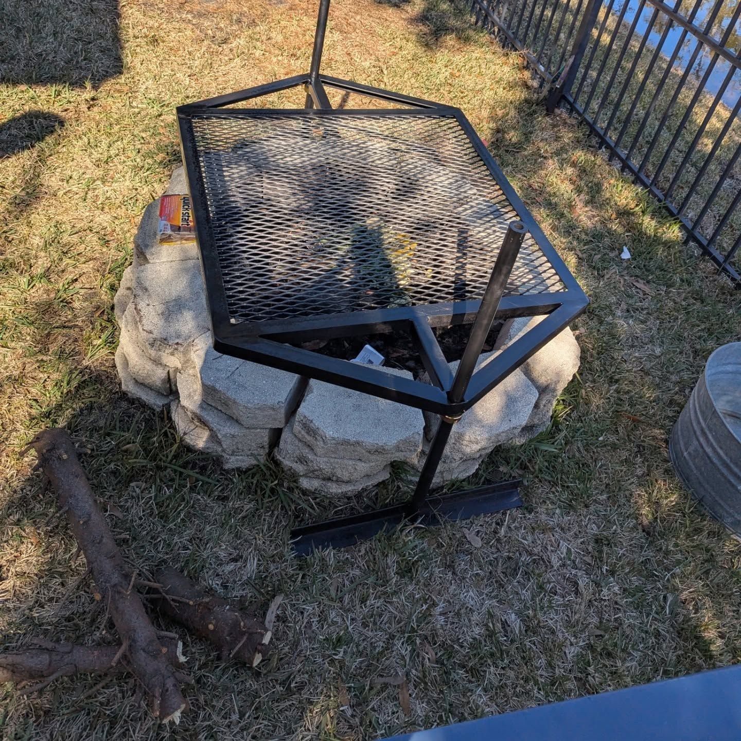 Fire pit with a metal grate and frame on a stone base, next to a log on grass.
