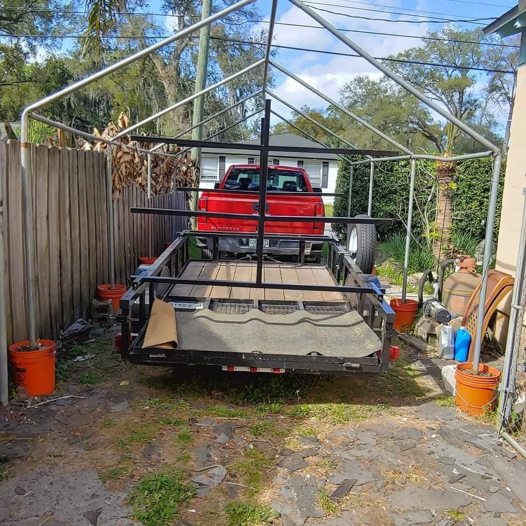 Red pickup truck on trailer under a metal canopy. Orange buckets flank the trailer; trees in background.