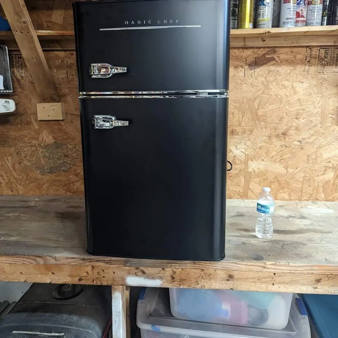 Black retro-style refrigerator on a wooden shelf in a garage, next to a water bottle.
