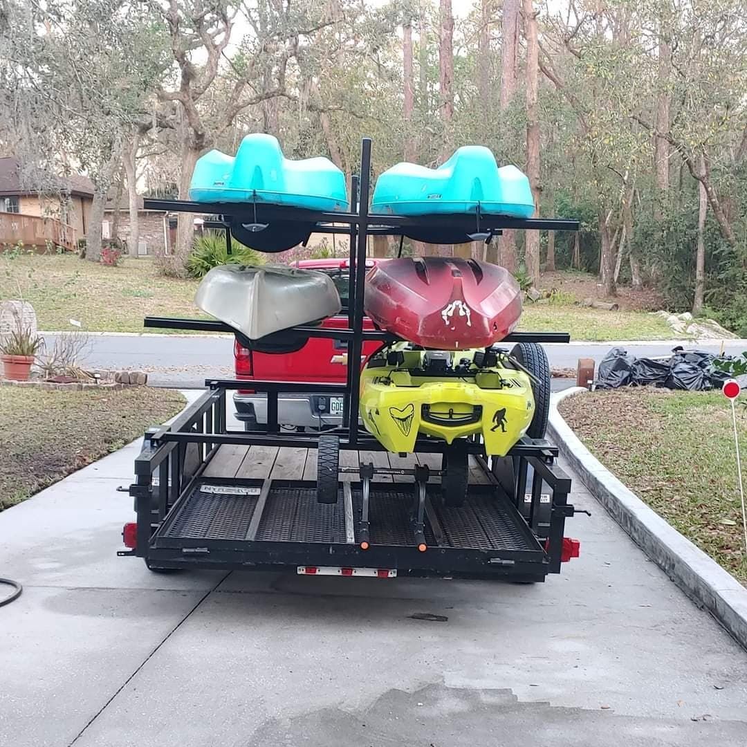 A black trailer with a rack holding four kayaks of various colors, on a driveway.